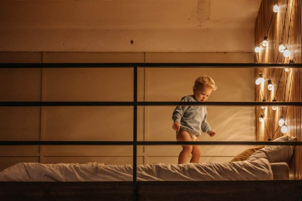 Charming toddler stands on bed in cozy bedroom with string lights adding a warm glow.