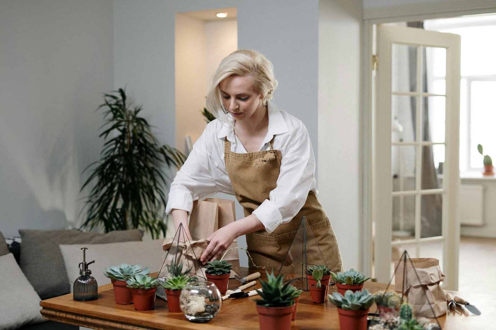 Blonde woman arranging succulent terrariums in a cozy home setting, wearing an apron.