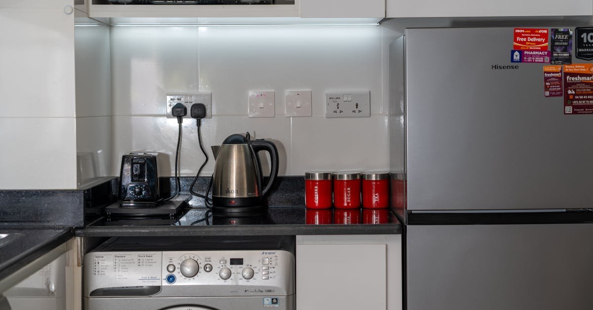 Stylish modern kitchen with stainless steel appliances and red accents, highlighting a washing machine, fridge, and countertop electronics.
