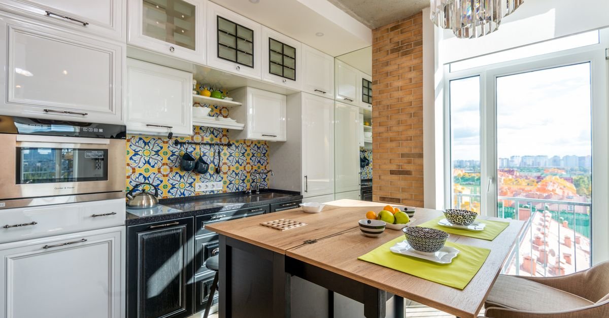 Chairs at wooden table with fruits placed near white cupboards and modern appliances in stylish kitchen with window at home
