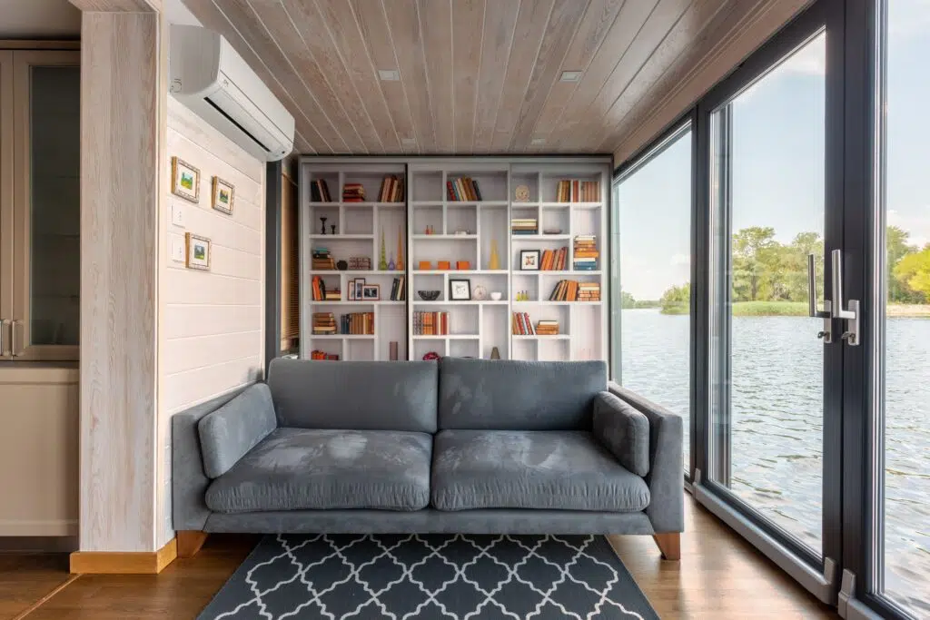 Interior design of modern house with majestic view on river surface through glass wall from comfortable soft gray couch placed in front of wide bookshelf
